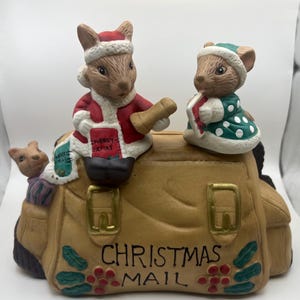 Puede incluir: Una bolsa de correo de Navidad de cerámica con tres figuritas de ratones. Los ratones llevan gorros de Papá Noel y llevan regalos. La bolsa es marrón con una hebilla dorada y las palabras "Christmas Mail" escritas en la parte delantera.