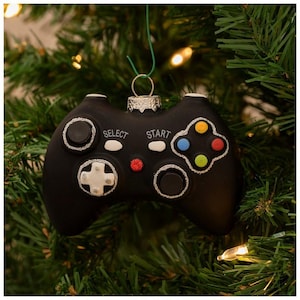 May include: Black glass Christmas ornament shaped like a video game controller with silver glitter accents. The buttons are colored red, blue, green, and yellow. The controller has the words "Select" and "Start" printed on it.