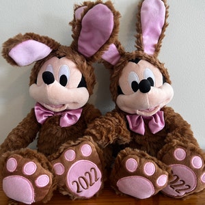 May include: Two brown plush Mickey Mouse toys wearing pink bunny ears and bow ties. Both toys have pink paw prints on their feet and the year 2022 embroidered on their paws.