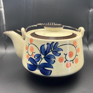 Könnte beinhalten: Eine cremefarbene Teekanne mit braunem Rand und einem floralen Design in Blau und Orange. Die Teekanne hat einen Metallgriff und einen Deckel.