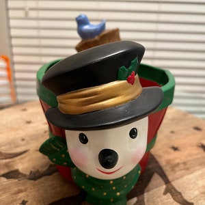 Könnte beinhalten: Ein Keramik-Schneemann-Pflanzgefäß mit schwarzem Hut, goldenem Rand und grünem Schal. Der Pflanztopf ist rot und grün mit einem kleinen blauen Vogel obenauf. Der Schneemann hat ein lächelndes Gesicht und ist mit Stechpalmen verziert.