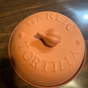 May include: A round, terracotta-colored ceramic container with a lid. The lid features the embossed text GARLIC and TORTILLA, arranged in a circular pattern, and is topped with a small, sculpted handle shaped like a single bulb of garlic.