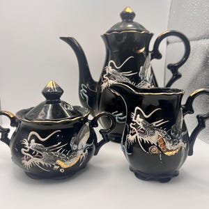 Op de afbeelding: Een zwart keramisch theeservies met een theepot, een melkkan en een suikerpot. Elk stuk is versierd met een gedetailleerd drakenontwerp in zilver, goud en wit. De set heeft gouden accenten en een glanzende afwerking.