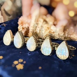 Könnte beinhalten: Nahaufnahme von fünf silbernen Ringen mit tropfenförmigen, irisierenden Mondstein-Cabochons. Die Mondsteine haben einen perlmuttartigen weißen und blauen Schimmer. Die Ringe liegen auf einer dunkelblauen Samtoberfläche.