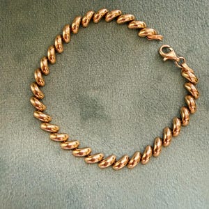 Puede incluir: Una pulsera dorada con una serie de eslabones redondeados y conectados. La pulsera se asegura con un cierre de mosquetón. La joya se muestra sobre una superficie verde.