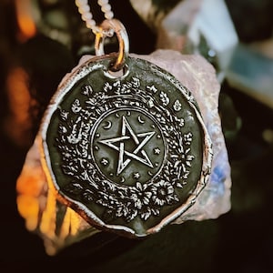 Puede incluir: Colgante plateado con un diseño detallado que presenta una estrella de cinco puntas, medias lunas y elementos florales. El colgante cuelga de una delicada cadena. El diseño está sobre un fondo oscuro.