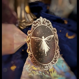 May include: An oval-shaped, ornate, gold-colored pin featuring a celestial fairy design. The fairy is depicted with outstretched wings against a dark background with small star-like details. The pin has a decorative, swirling border.