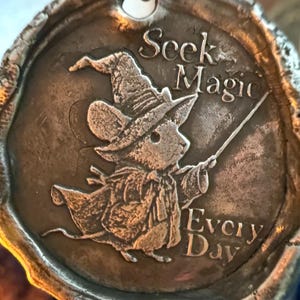May include: A silver-toned pendant with a raised image of a mouse dressed as a wizard, holding a wand. The pendant is circular with the words "Seek Magic Every Day" embossed around the mouse.