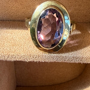 Op de afbeelding: Een gouden ring met een grote, ovaal geslepen, gefacetteerde paarse edelsteen in een gladde, gepolijste gouden zetting. De ring heeft een eenvoudige, massief gouden band die aansluit op de zijkanten van de ovale zetting met een glanzende afwerking.