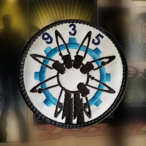 May include: A circular embroidered patch featuring a white background with a central blue gear design. Overlaid are black silhouettes of five knives arranged in a starburst pattern. The numbers 9, 3, and 5 are embroidered in blue at the top.