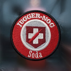 May include: Circular embroidered patch featuring the text "JUGGER-NOG" in white, arched at the top, and "Soda" at the bottom. The design includes a white cross on a grey background, with a red syringe. The patch has a red border with a black edge.