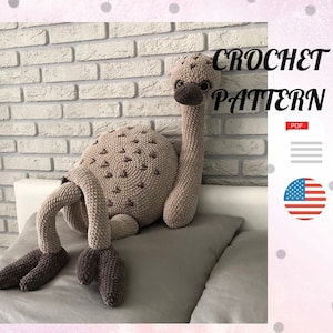 May include: Crochet pattern for a brown and beige amigurumi ostrich. The pattern is available as a PDF download. The image shows the finished amigurumi ostrich lying on a bed.