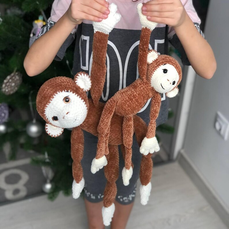 May include: Two brown crocheted monkey toys with white faces and white hands hanging from a person's hands. The monkeys are made of soft, fuzzy yarn.