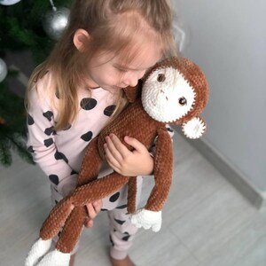 May include: A young girl is holding a brown and white crocheted monkey plush toy. The monkey has a long tail and white fur on its hands and feet.