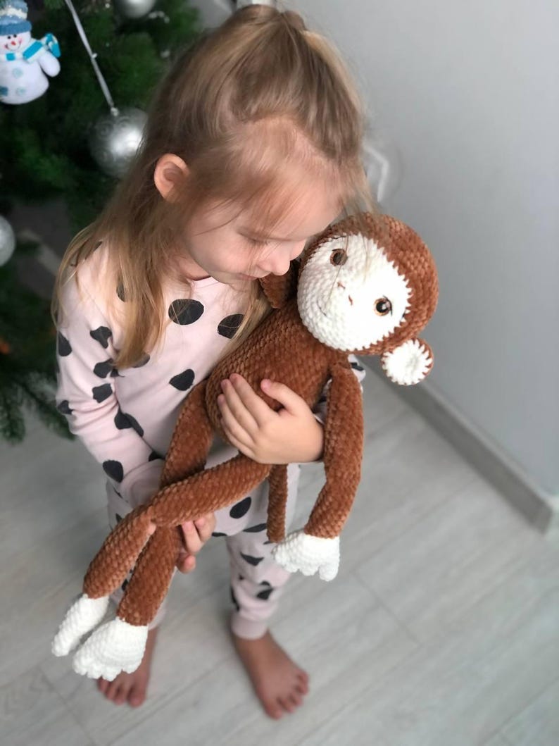 May include: A young girl is holding a brown and white crocheted monkey plush toy. The monkey has a long tail and white fur on its hands and feet.