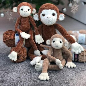 May include: Three crocheted monkey toys, one brown, one beige, and one smaller beige, sitting on a gray fuzzy surface. The larger brown monkey has white eyes and a white face.