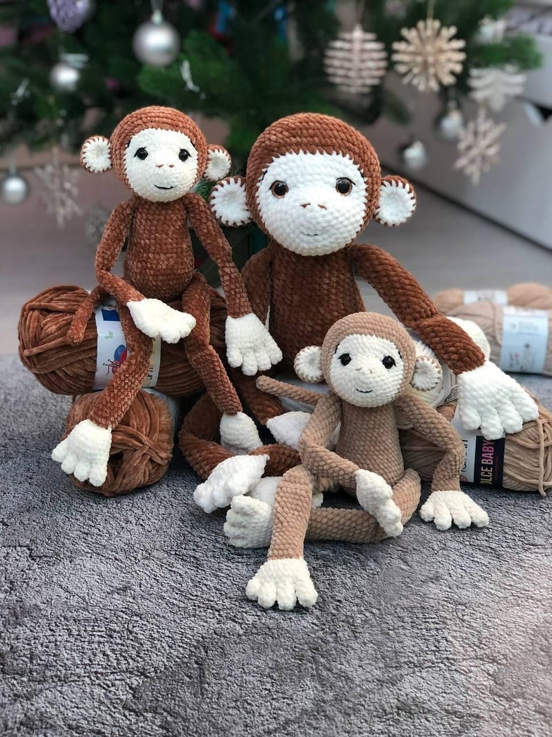 May include: Three crocheted monkey toys, one brown, one beige, and one smaller beige, sitting on a gray fuzzy surface. The larger brown monkey has white eyes and a white face.