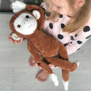 May include: A brown and white crocheted monkey toy with a long tail. The monkey has a white face with brown eyes and a brown nose. The monkey is being held by a child.
