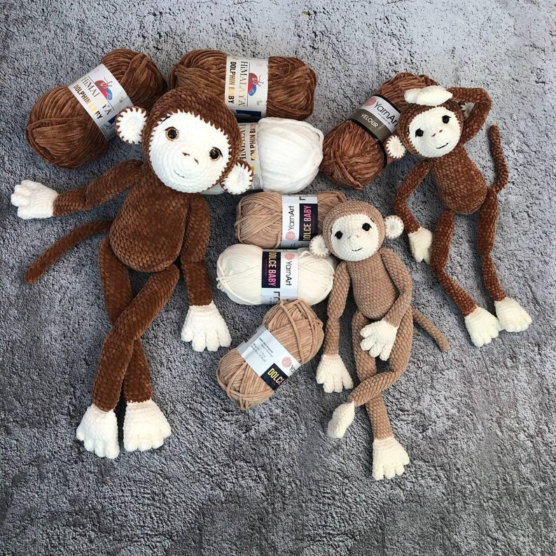 May include: Three brown crocheted monkey toys with white faces and yarn balls in various colors, including brown, beige, and white. The yarn balls have labels with the text "YarnArt Dolce Baby" and "HIMALAYA DOLPHINE BY".