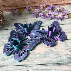 May include: Three iridescent butterfly-shaped candle holders. The candle holders are a gradient of blue and purple with a teal-colored candle cup. The butterfly wings have intricate cut-out designs. The background includes purple flowers.