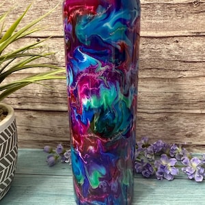 May include: A tall, insulated tumbler with a clear lid. The tumbler features a vibrant, swirling design in shades of blue, purple, pink, and green, creating a marbled effect. The design is accented with subtle glitter. The tumbler is designed to keep drinks cold or hot.