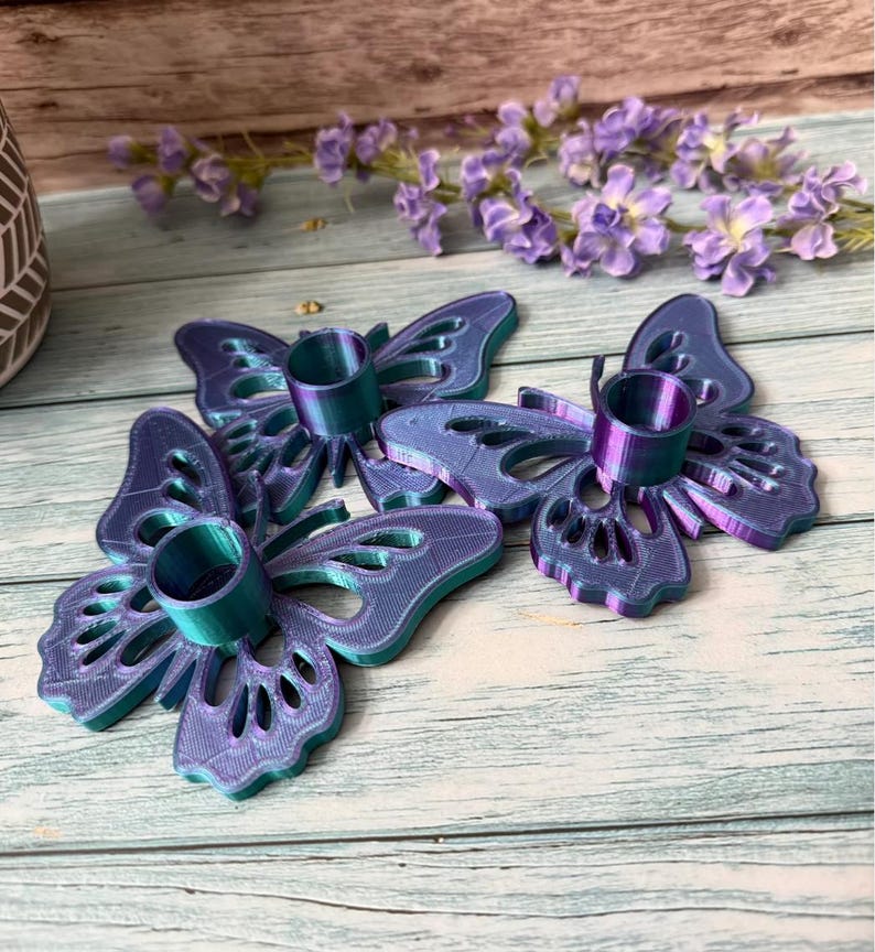 May include: Three iridescent butterfly-shaped candle holders. The candle holders are a gradient of blue and purple with a teal-colored candle cup. The butterfly wings have intricate cut-out designs. The background includes purple flowers.
