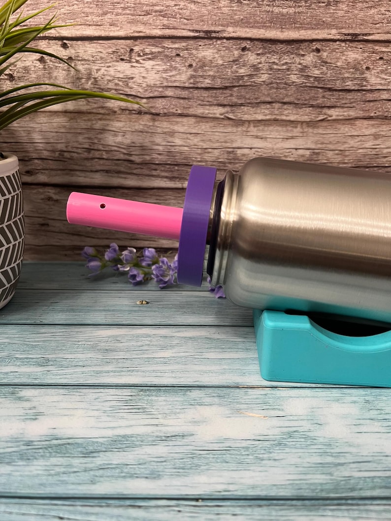 Peut inclure: Une bouteille d'eau en acier inoxydable avec une paille rose et un couvercle violet. La bouteille est pos&eacute;e sur un support turquoise.