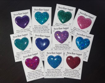 May include: A collection of heart-shaped glitter resin pocket hugs in various colors, including purple, teal, blue, pink, and red. Each heart is on a white card with the text "You've Been Hugged" and a message.