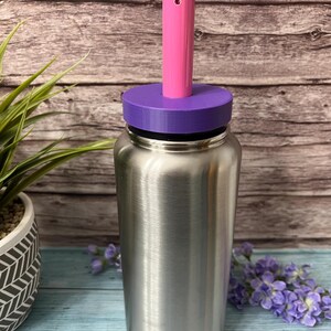 Peut inclure: Une bouteille d'eau en acier inoxydable avec un couvercle violet et une paille rose. La bouteille est pos&eacute;e sur une surface en bois.