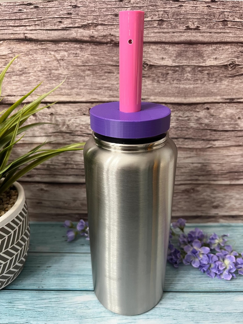 Peut inclure: Une bouteille d'eau en acier inoxydable avec un couvercle violet et une paille rose. La bouteille est pos&eacute;e sur une surface en bois.
