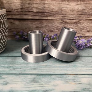 Peut inclure: Deux bougeoirs en m&eacute;tal argent&eacute; avec une base ronde et un dessus cylindrique. Les bougeoirs sont pos&eacute;s sur une surface en bois bleu clair.