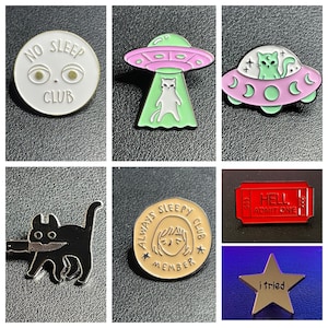 May include: A set of nine enamel pins featuring various designs, including a cat with a knife, a cat in a UFO, a cat in a spaceship, a ticket that says "Hell, Admit One", a star with the text "I tried", and a round pin with the text "Always Sleepy Club Member".