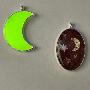 May include: Two silver charms, one shaped like a crescent moon with a bright green fill, the other an oval with a red glitter fill and a white crescent moon and stars.