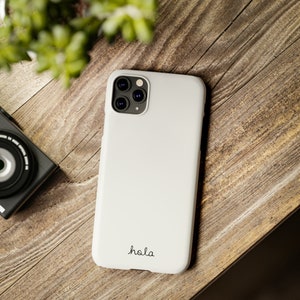 May include: White iPhone case with a black camera lens and the word "hola" written in black cursive.