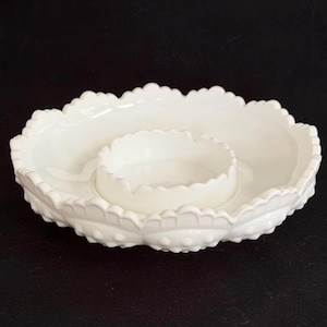 May include: A white, oval-shaped serving dish with a central, smaller bowl. The outer rim has a scalloped edge with a textured, leaf-like pattern. The dish is made of ceramic or porcelain.