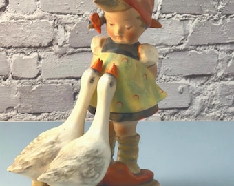 May include: A ceramic figurine of a child wearing a red headscarf and a yellow dress with a dark bodice, standing on a base with two white geese. The geese have orange beaks and feet, and the child has light brown hair and is looking downward.