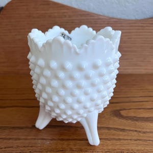 Könnte beinhalten: Eine weiße Hobnail-Glasvase mit drei Beinen. Die Vase hat einen gewellten Rand und eine strukturierte Oberfläche, die mit kleinen, abgerundeten Noppen bedeckt ist. Die Vase steht auf einer Holzoberfläche.