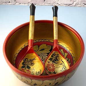 May include: A large round wooden bowl with a red exterior and gold interior, featuring hand-painted black and red floral patterns. Two matching wooden spoons with black handles and gold-painted bowls rest inside, displaying similar floral motifs.