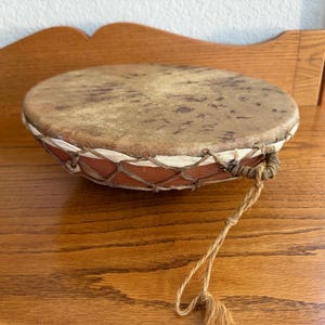 May include: A small, round drum with a natural hide drumhead. The drum's body is a reddish-brown color, with a woven net design around the sides. A natural fiber cord is attached to the side of the drum.