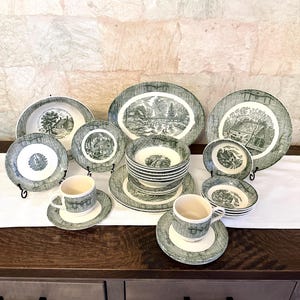 May include: A collection of ceramic dinnerware featuring a dark green and white transferware pattern of rural landscapes. The set includes various plates, bowls, and cups with saucers, all decorated with detailed scenes of houses and trees.