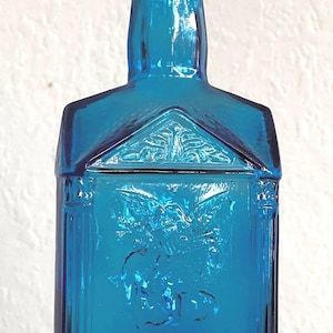 Puede incluir: Una vibrante botella de vidrio azul con un diseño arquitectónico único. La botella presenta detalles en relieve, incluyendo un águila y texto. La botella tiene forma rectangular con una parte superior puntiaguda y está hecha de vidrio transparente.