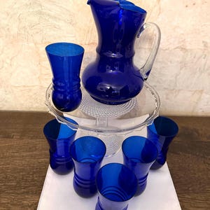 May include: A cobalt blue glass pitcher with a clear glass handle and six matching cobalt blue glass tumblers. Each tumbler features a rounded base with three horizontal ridges. The set is arranged on a clear glass pedestal stand.