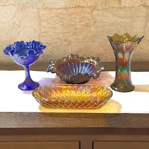 May include: A collection of four iridescent glass pieces: a blue ruffled compote, a dark purple ruffled bowl, a tall green scalloped vase, and an amber oval dish with a diamond-patterned exterior. Each item features a reflective finish.