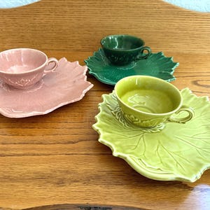May include: Three ceramic tea cups and matching saucers in pink, dark green, and light green. Each saucer is shaped like a large leaf with detailed veining, and each cup features a matching embossed leaf pattern at the base and a curved handle.