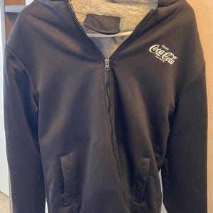 May include: Black zip-up hooded jacket with a grey faux fur lining. The jacket features the Coca-Cola logo in white on the upper chest. The jacket has a ribbed hem and cuffs and a hood.