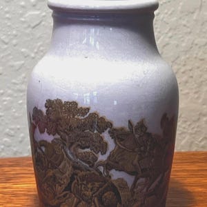 May include: A light purple vase with a detailed brown relief design depicting a hunting scene with figures on horseback, trees, and animals. The vase has a slightly flared rim and a smooth, glossy finish.