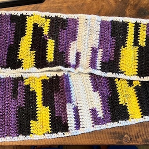 May include: A handmade crochet scarf with a repeating pattern of yellow, black, purple, and white vertical stripes. The scarf is made with a soft yarn and has a textured appearance. The scarf is approximately 178 cm long.