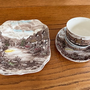Könnte beinhalten: Ein Satz von vier unregelmäßig geformten Tellern und eine Teetasse mit Untertasse. Die Teller und die Untertasse zeigen eine braune und weiße Landschaftsszene. Die Teetasse ist innen weiß.