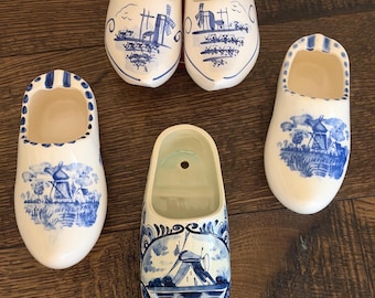 May include: Five ceramic clogs featuring blue and white hand-painted designs of windmills and rural landscapes. Each shoe-shaped ornament has a hollow interior and a smooth, glossy finish, with some pieces displaying intricate floral patterns.