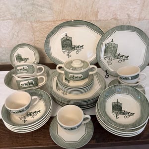 May include: A collection of cream-colored ceramic dinnerware featuring a dark green border with a textured pattern and a central illustration of a horse-drawn carriage in front of a building. The set includes plates, bowls, cups, and a lidded tureen.
